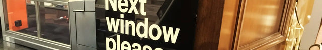 Black acrylic counter sign with white text reading 'Next window please' at a service desk