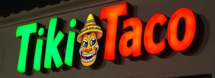 Illuminated storefront channel letter sign reading 'Tiki Taco' in green and red letters with a cartoon face wearing a sombrero between the words, against a dark background
