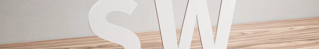 Flat cut white acrylic letters SW standing on a light wood surface against a neutral background