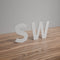 Flat cut acrylic letters SW in white, displayed on a wooden surface against a plain wall