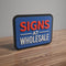 Multi-layered 3D sign with bold red and white letters reading 'Signs at Wholesale' on a blue background, placed on a wooden surface
