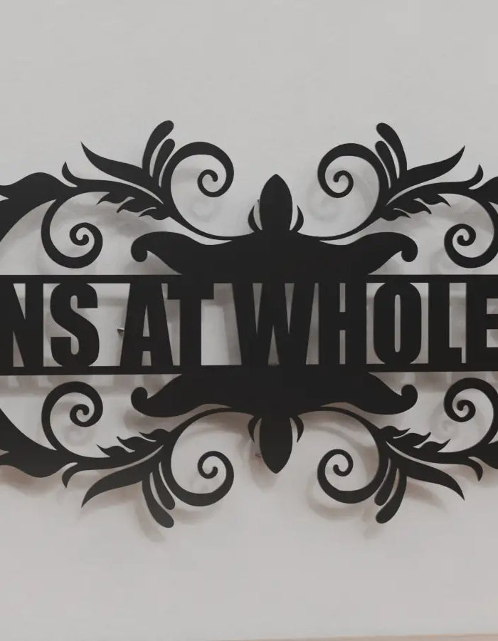 metal monogram sign displaying 'SIGNS AT WHOLESALE' in a laser-cut black design with decorative scrollwork, mounted on a plain light wall above a wooden surface.