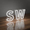 On Static LED neon signs letters SW glowing in white, displayed on a wooden surface with reflections against a gray background