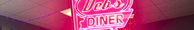 Uv-printed Neon sign reading 'Deb's Diner' in bright pink letters with an arrow, hanging indoors against a ceiling background