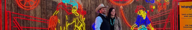 Colorful neon cowboy-themed display with Western motifs, including rodeo riders, horses, boots, and Wrangler signage, featuring two people posing in front